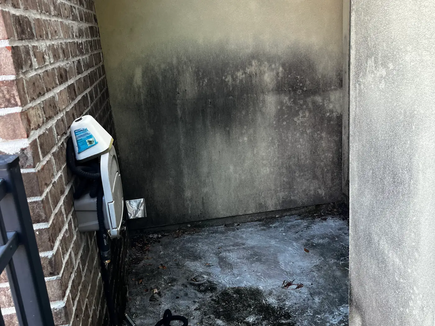 A stucco wall covered in heavy black mildew with a filthy concrete pad before soft washing.